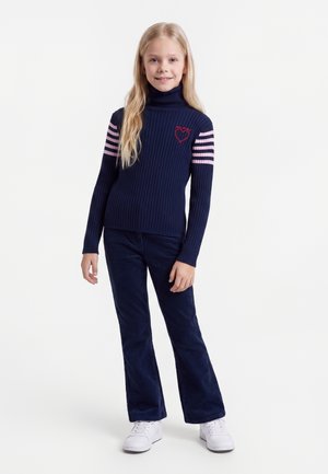 Ung pige med langt blondt hår, iført en marineblå rullekravesweater med pink striber og hjerte-detalje, marineblå bukser og hvide sneakers.