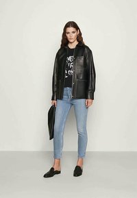 Jaqueta de couro preta com uma textura suave, apresentando um fecho com zíper e dois bolsos frontais. Combinada com uma t-shirt gráfica e jeans azul claro.