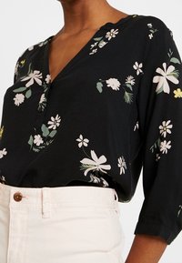 Blouse florale noire avec un décolleté en V, des manches trois quarts et une coupe décontractée. Présente des motifs de fleurs blanches et vertes sur un fond sombre.
