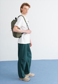 Mochila verde sobre una persona de pie que lleva una camiseta blanca y pantalones anchos verdes, con zapatillas beiges sobre un suelo azul.