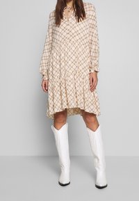 Light beige long-sleeve dress with a checkered pattern in brown tones, featuring a tiered hem and a tie detail at the neckline. Paired with white boots.