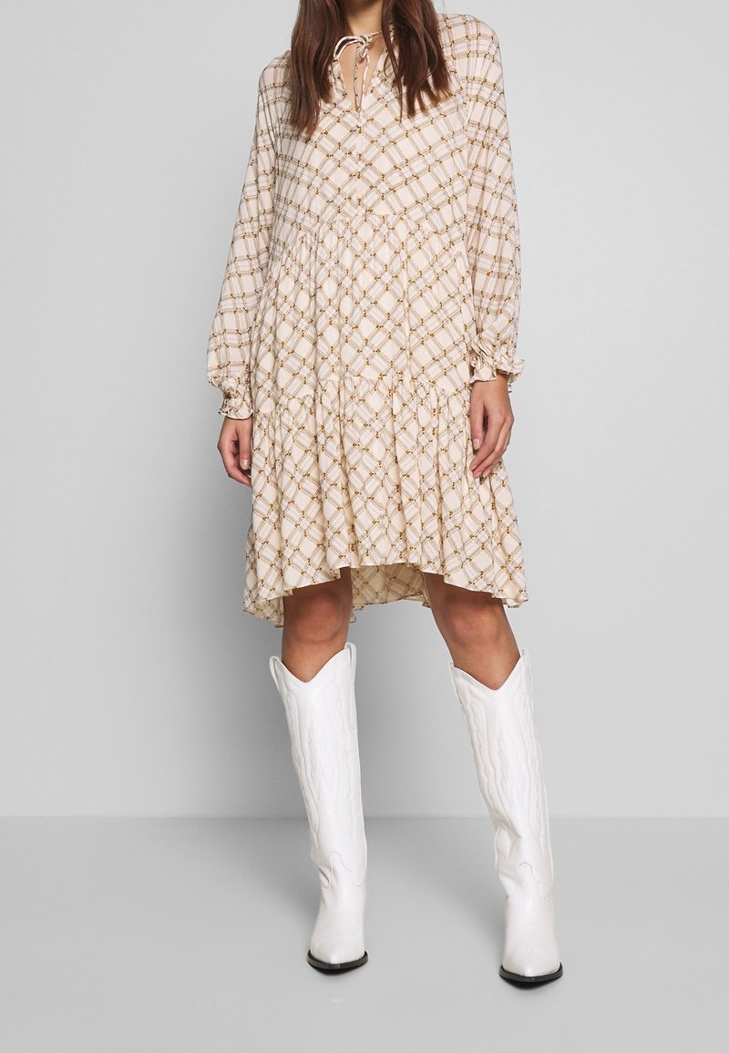 Light beige long-sleeve dress with a checkered pattern in brown tones, featuring a tiered hem and a tie detail at the neckline. Paired with white boots.
