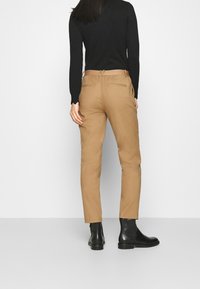 Pantalons beiges en tissu lisse avec une coupe ajustée, dotés de deux poches arrière. Portés avec des bottines noires et un haut à manches longues noir.
