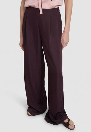 Vrouw draagt donkerpaarse, wijde broek met hoge taille, een lichtroze blouse en bruine open hakken, staand voor een effen achtergrond.