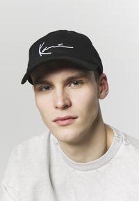 Svart keps med vit broderad signatur på framsidan, böjd skärm och en texturerad tyg. Modellen bär en ljusgrå sweatshirt.