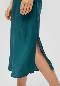Turkusowa sukienka z lekkiego materiału, z bocznym rozcięciem i zaokrągloną dolną krawędzią. Subtelna faktura i mały metalowy akcent w pobliżu dolnej krawędzi.