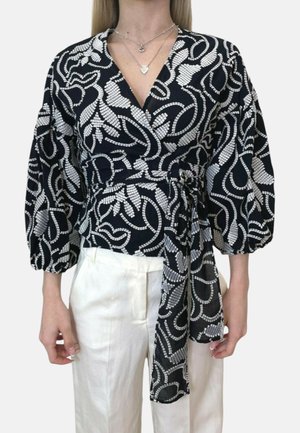 Donna che indossa una blusa nera a portafoglio con motivo astratto bianco, maniche a sbuffo e cintura in vita, abbinata a pantaloni bianchi a vita alta.