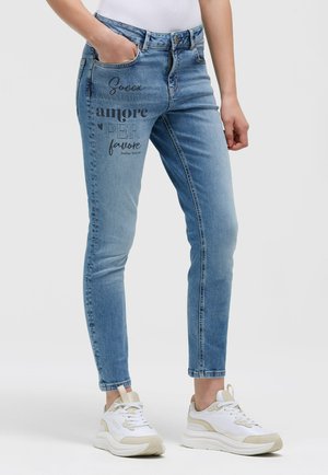 Light blue slim-fit jeans with black printed text on the left thigh, worn with white and beige sneakers and a white top.