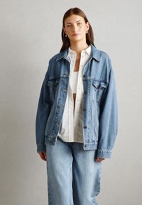 Giubbotto in denim azzurro chiaro con vestibilità oversize, tasche frontali e bottoni marroni. Indossato sopra una camicia bianca, abbinato a jeans a gamba larga azzurro chiaro.