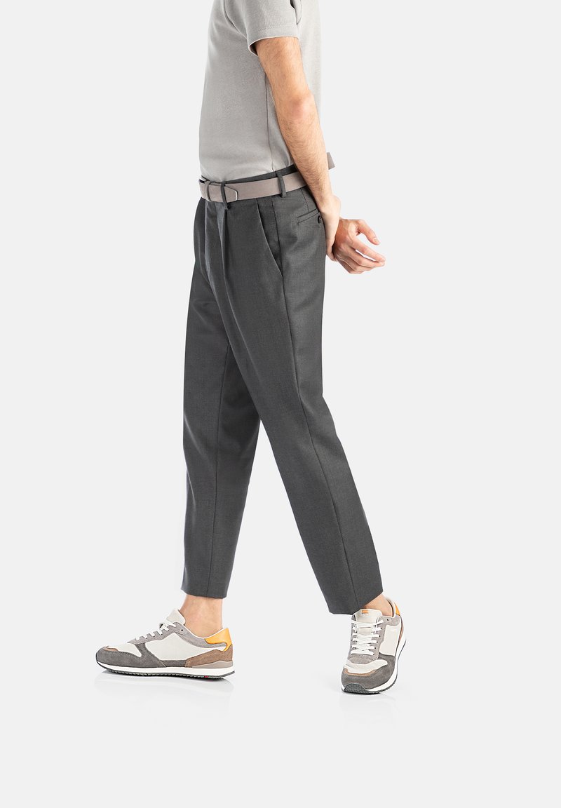Graue Hose mit Vorderfalten und lockerer Passform, kombiniert mit beigen und grauen Sneakers mit einem orangefarbenen Akzent. Hellgraues Hemd.