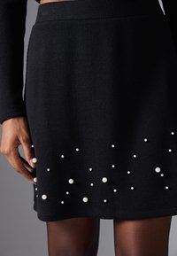 Jupe noire à côtes avec des accents de perles blanches dans un motif éparpillé. Design mi-long avec une ceinture ajustée et un tissu texturé.