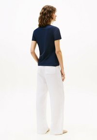 Mujer con cabello rizado vistiendo una camiseta ajustada de color azul marino, pantalones blancos holgados con bolsillos traseros y zapatos de beige trenzados, de pie de lado.