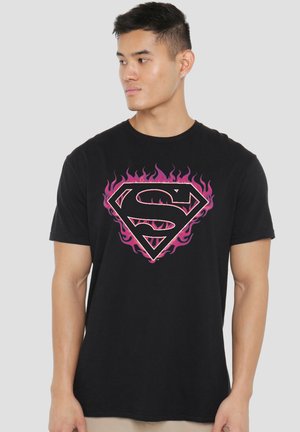 Junger Mann trägt ein schwarzes T-Shirt mit einem flammenden pinken Superman-Logo und schaut nach links vor einem schlichten hellen Hintergrund.