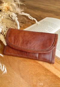 Brown leather wallet with rounded flap, stitched edges, and a smooth texture. Placed on a wooden surface near dried flowers and a book.