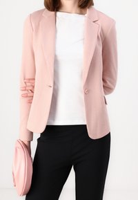 Světle růžový blazer s přiléhavým střihem, zapínání na jeden knoflík a klopovými límci; nošený přes bílý top a kombinovaný s černými kalhotami.