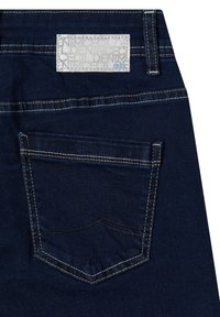 Los vaqueros de mezclilla azul oscuro presentan una etiqueta rectangular blanca con texto en relieve cosida en la cinturilla, y un bolsillo trasero con costuras en contraste.