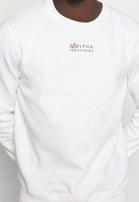 Felpa bianca in tessuto morbido, con collo tondo e logo "Alpha Industries" ricamato in beige sul petto.