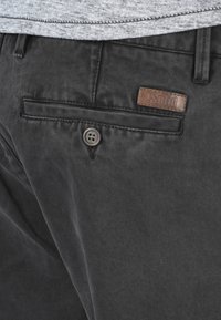Pantalons en tissu noir avec fermeture à bouton et une étiquette en cuir sur la poche arrière, avec une texture lisse et une coupe ajustée.