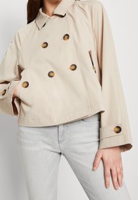 Chaqueta beige corta con un diseño de doble botonadura, grandes botones oscuros y mangas anchas. Combinada con unos pantalones vaqueros gris claro.