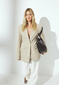 Beige übergroßer Blazer mit langen Ärmeln, frontalen Taschen und tiefem V-Ausschnitt, kombiniert mit weißen weiten Hosen und einer schwarzen Ledertasche.