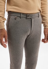 Pantaloni grigi su misura realizzati in tessuto testurizzato. Presentano una parte frontale liscia, due bottoni e tasche laterali. Indossati con un maglione beige.
