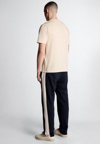 Beige katoenen t-shirt en marineblauwe joggingbroek met off-white zijstrepen. Model gezien van achteren, met lichtgekleurde schoenen.