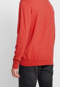 Pull en maille rouge à manches longues, col rond et ourlet côtelé. Texture douce, coupe décontractée, porté avec un jean gris foncé.