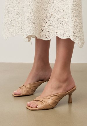 Lábak bézs szandálban, csavart pántokkal és négyzet alakú orr-résszel, sima padlón állnak, részben fehér horgolt szoknyával takarva.