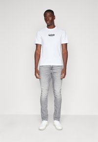 Hvit bomulls t-skjorte med svart logo, kombinert med lysegrå slim-fit bukser og hvite sneakers, stående mot en ensfarget bakgrunn.