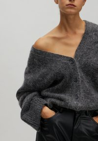 Grijze wollen cardigan met een V-hals, voorzien van drie zwarte knopen en brede mouwen. Gedragen over zwart leren broek, met een gestructureerde stof.