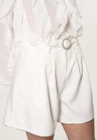 Pantaloni corti bianchi a vita alta con pieghe, caratterizzati da una fibbia per cintura rotonda e decorata e una texture liscia. L'orlo presenta dettagli di rifinitura discreti.