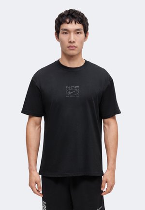 Νέος άνδρας που φοράει μαύρο μπλουζάκι Nike Athletic Club και μαύρο σορτς, στέκεται στραμμένος προς τα εμπρός μπροστά σε ένα απλό φωτεινό φόντο.