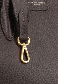 Bruin leren tas met een textuur, voorzien van een gouden metalen sluiting en stiksel details. Logo label zichtbaar aan de bovenkant in gouden letters.
