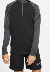 Svart sporttop med grå detaljer, halvkedja, lång ärm och nätmaterial på höger axel. Känns bra med svarta shorts som har en logotyp.