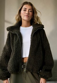 Dunkelgrüne Fleecejacke mit hohem Kragen, Reißverschluss und oversized Passform. Der Stoff hat ein strukturiertes, flauschiges Aussehen.