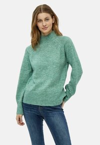Maglione lavorato a maglia verde menta con collo alto, orlo a coste e maniche lunghe, abbinato a jeans blu scuro. Texture morbida e vestibilità casual.