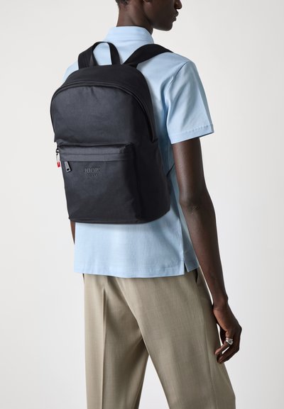 Sac à dos en tissu noir avec fermeture éclair et poche avant. Comprend des bretelles ajustables, un logo, et un design simple et épuré.