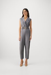 Grå langermet jumpsuit uten ermer med belte i livet, to knapper foran, tapered ben og glatt tekstur; kombinert med beige hæler.