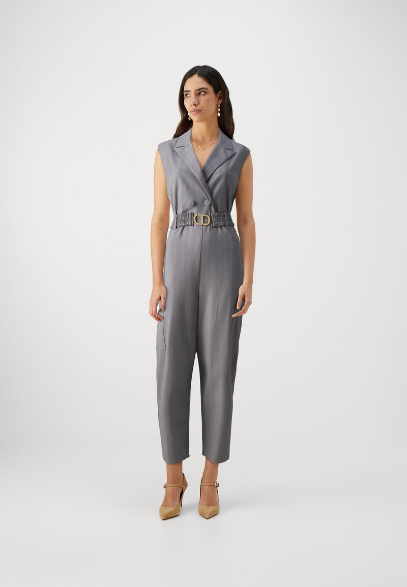Grå langermet jumpsuit uten ermer med belte i livet, to knapper foran, tapered ben og glatt tekstur; kombinert med beige hæler.