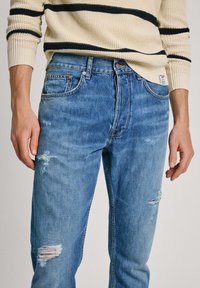 Vaalean siniset farkut, joissa on kulutettuja yksityiskohtia ja rento malli. Yhdistettynä kermaiseen neuleeseen, jossa on tummansinisiä raitoja hihoissa.