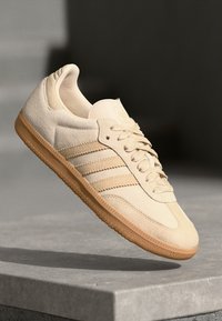 Bež suede tenisice s tri dijagonalne pruge, teksturirani gumeni potplat i zaobljenim vrhom. Lagani dizajn, minimalistički izgled.