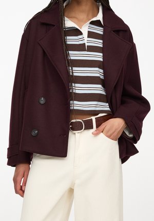 Mujer con abrigo corto burdeos, camiseta polo a rayas marrón y blanca, pantalones blancos con cinturón marrón, mano en el bolsillo.