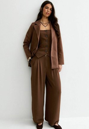 Donna che indossa un blazer marrone, un gilet abbottonato, pantaloni a gamba larga, ballerine nere e collane a strati, in piedi contro una parete bianca.