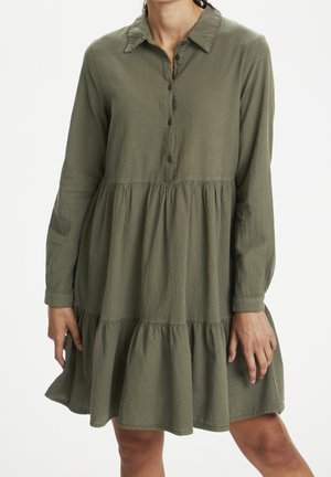 Femme portant une robe vert olive à manches longues, col, boutonnée sur le devant et une jupe à volants.