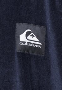 Jūros mėlynumo audinys su tekstūruota paviršiumi, kuriame yra stačiakampis juodas audinio pleistras, ant kurio pavaizduota balta banga bei užrašas "Quiksilver."