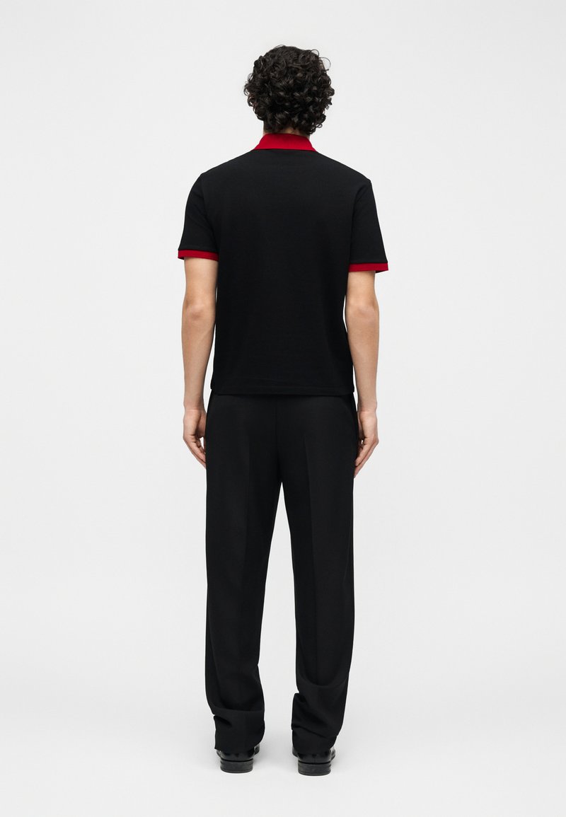 Man with curly hair stands facing away wearing black polo shirt with red collar and cuffs, black pants, and black shoes on white background.