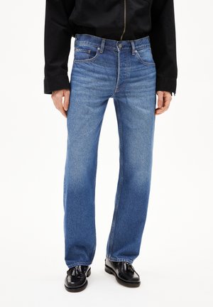 Mannelijke model draagt rechte blauwe jeans in combinatie met zwarte tassel loafers en een zwart long-sleeve top, staand tegen een witte achtergrond.