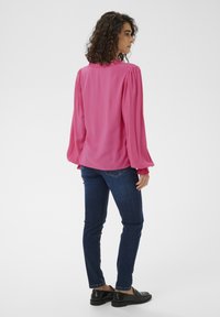 Blouse rose à manches bouffantes et col à volants, associée à un jean bleu foncé et des chaussures slip-on noires. Le tissu semble lisse et léger.