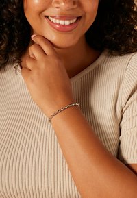 Zilveren armband met kleine ronde kralen, gedragen om een lichtbruine pols. De drager heeft krullend haar en draagt een neutrale, ribgebreide beige top.