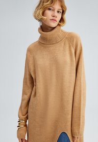 Beige turtleneck sweater med ribbet krave, lange ærmer og sidesslids. Blød tekstur og afslappet pasform med kontrasterende denim nedenfor.
